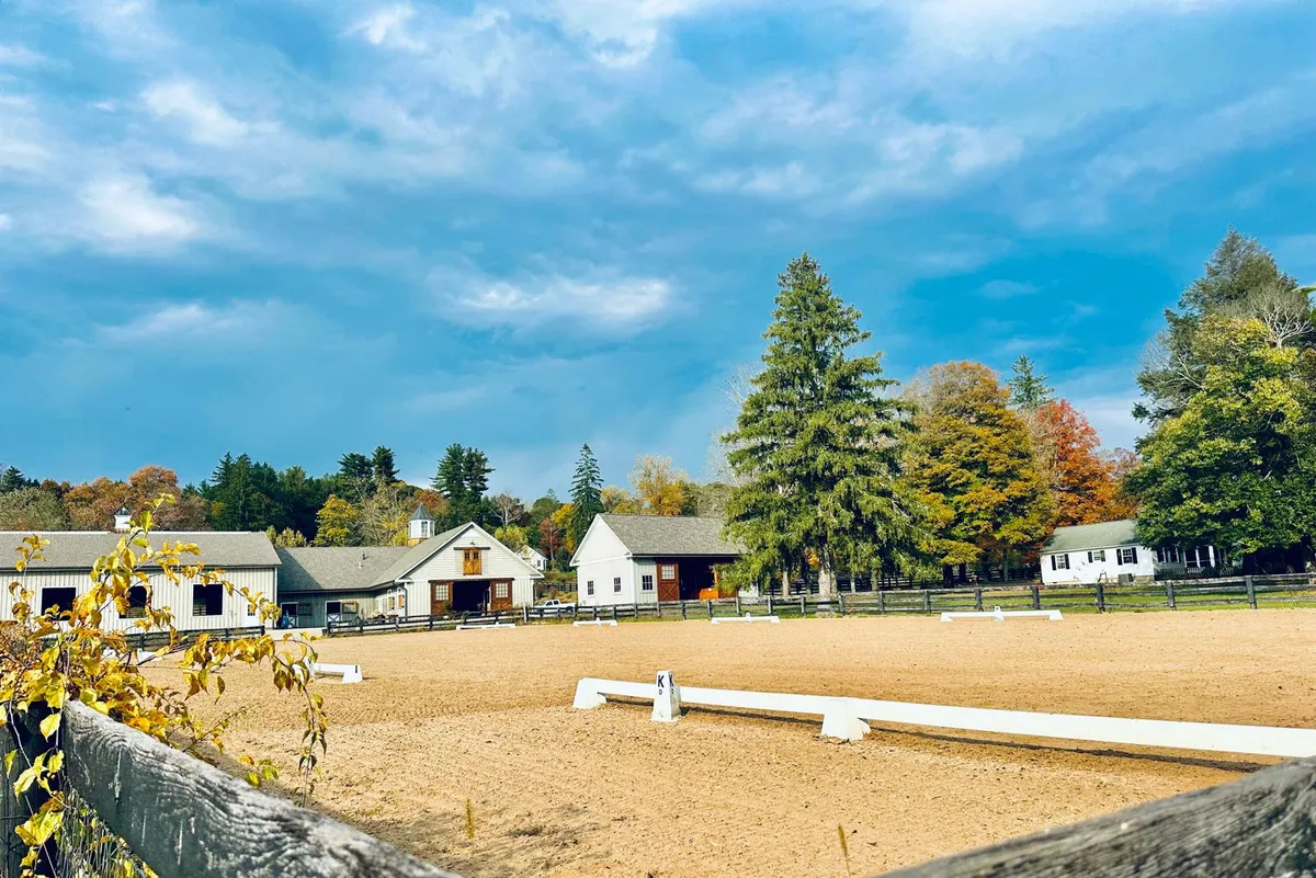 The outdoor sand arena with white fencing and stables in the autumn background